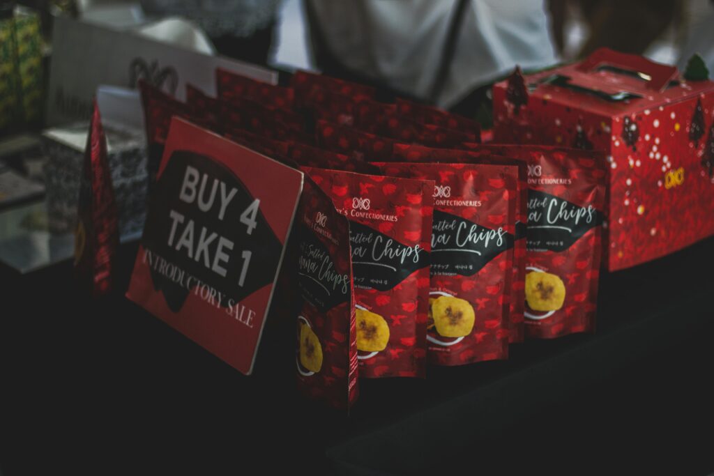 Packaged banana chips displayed with a promotional 'Buy 4 Take 1' sign at an indoor sale event.