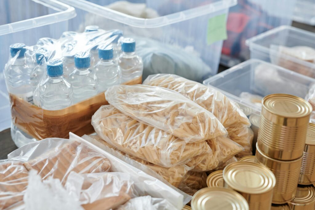 Plastic-wrapped food items and bottled water organized in storage containers and tin cans.